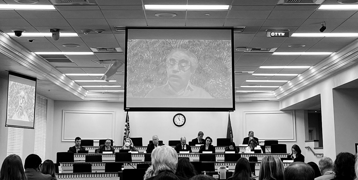 A room with a few dozen people.  Above, a big screen with an image of somebody speaking.