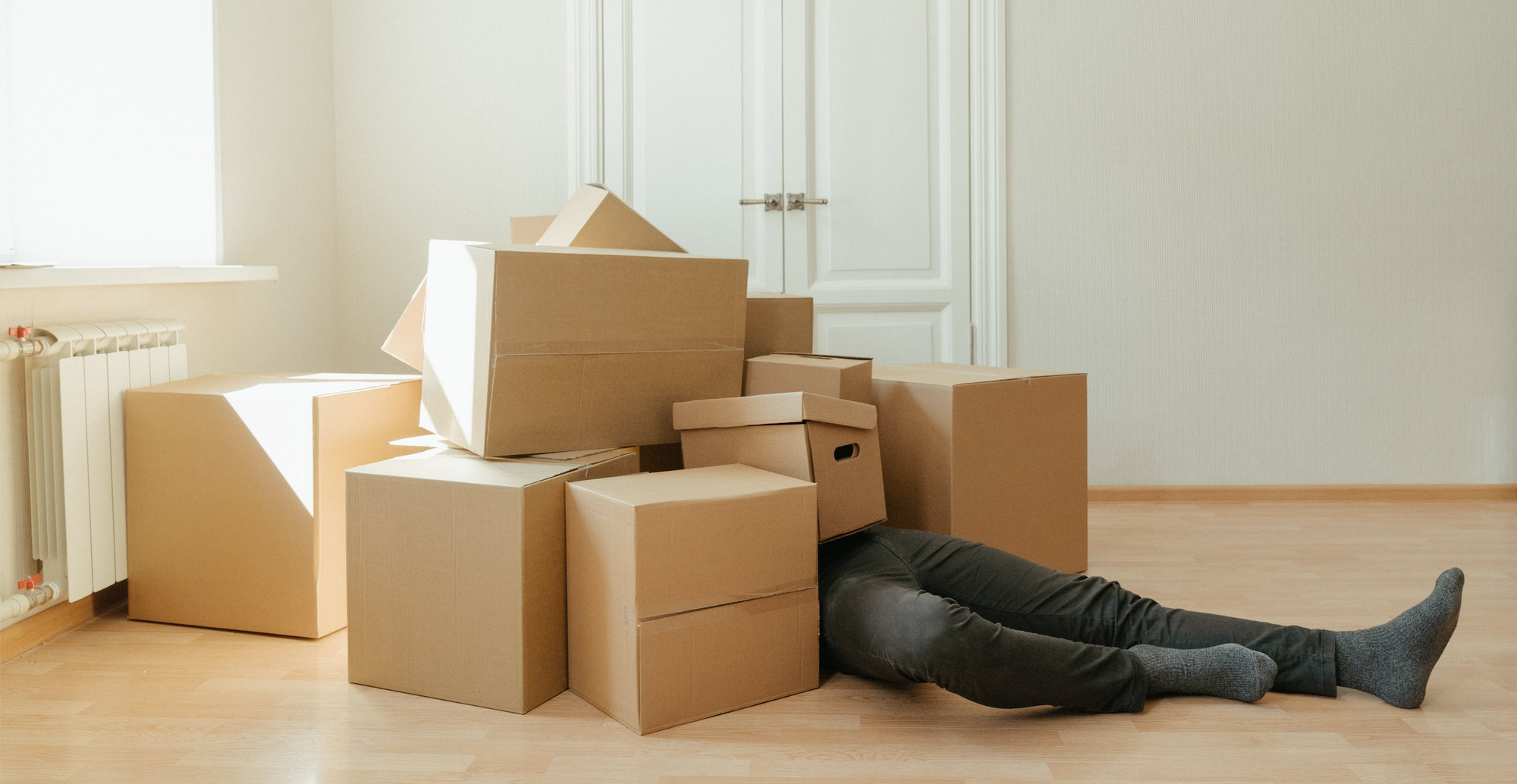Brown cardboard moving boxes on the floor, with a person beneath them whose feet are sticking out