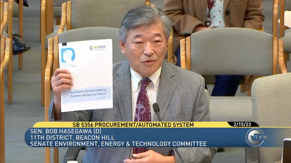 A Japanese man, in a tie and jacket, holding up paper.  Below, the words "SB 5356 Procurement/Automated System"